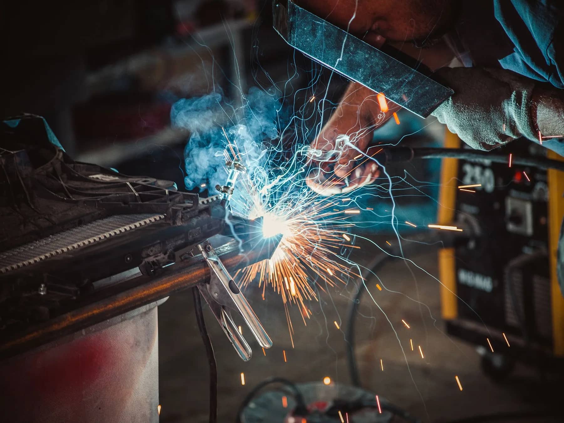 Welder laying a bead on a steel part, with a ground clamp on the workpiece and sparks flying